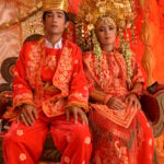 Para nowożeńców. Mężczyzna i kobieta mają na sobie bogato zdobione, czerwone stroje. Mają bogate, złote nakrycia głowy.