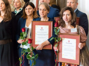 Zbliżenie na kobiety stojące w pierwszym rzędzie. Dwie z nich trzymają w dłoniach dyplomy w ramach i róże.